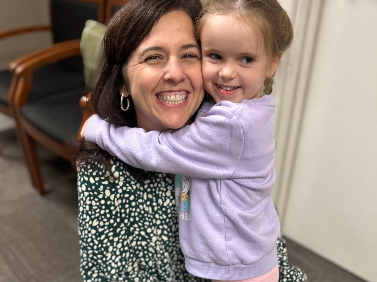 Occupational therapist smiling as she hugs a happy young child in a purple sweater at the New Heights Therapy clinic
