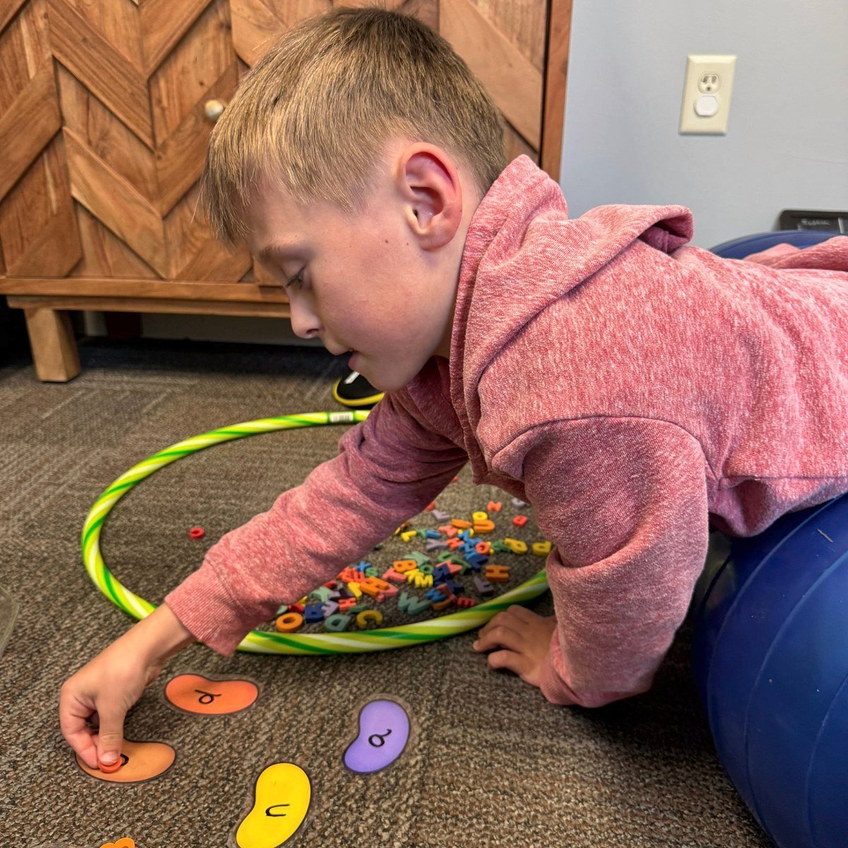 Young child in a pink hoodie reaching for letter tiles from a green sensory loop while draped over a therapy ball — building fine motor and motor planning skills during pediatric occupational therapy at New Heights Therapy in Kansas