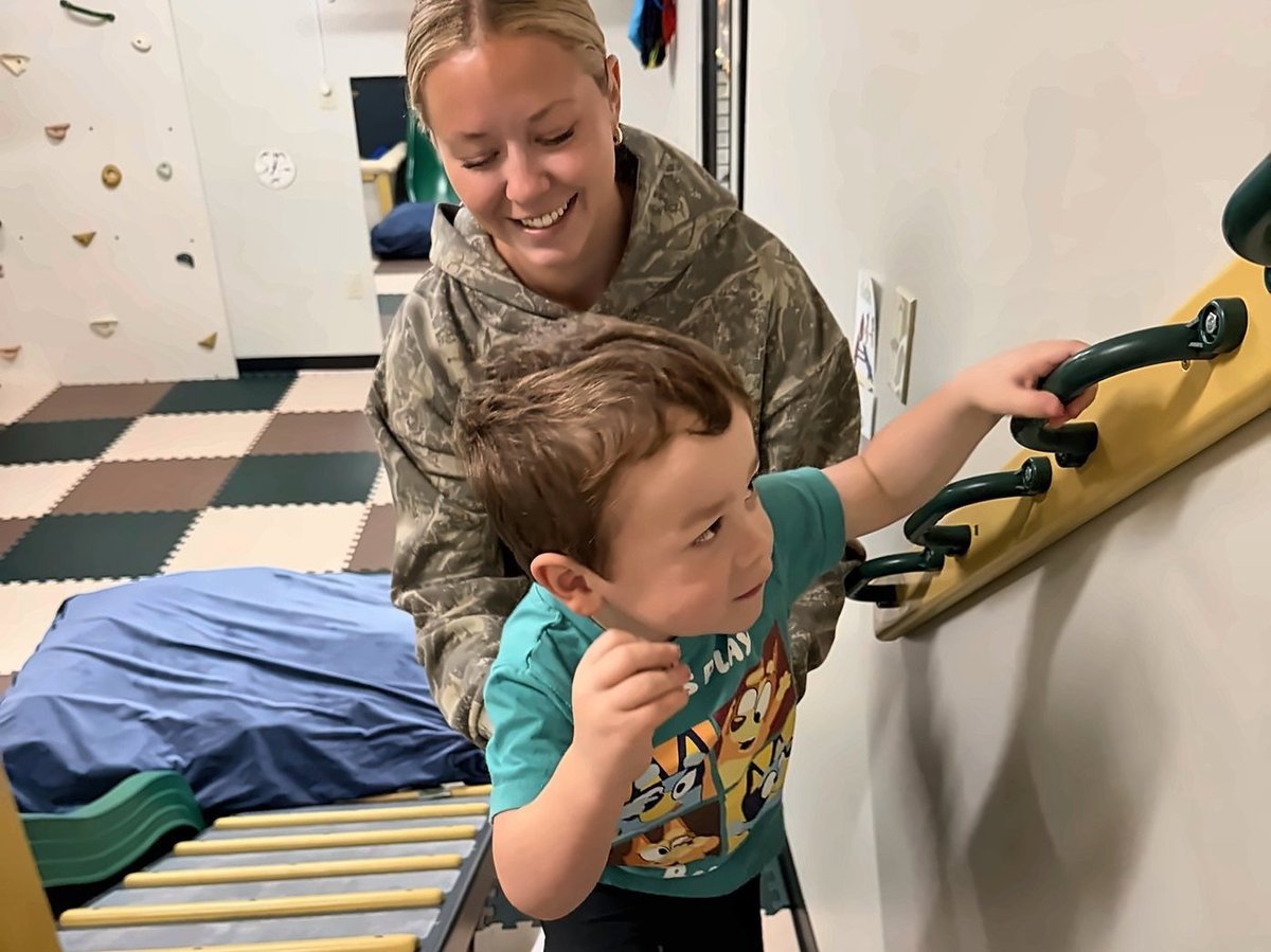 Toddler climbing stairs with a handrail during pediatric physical therapy, a therapist close by as he reaches for each handle