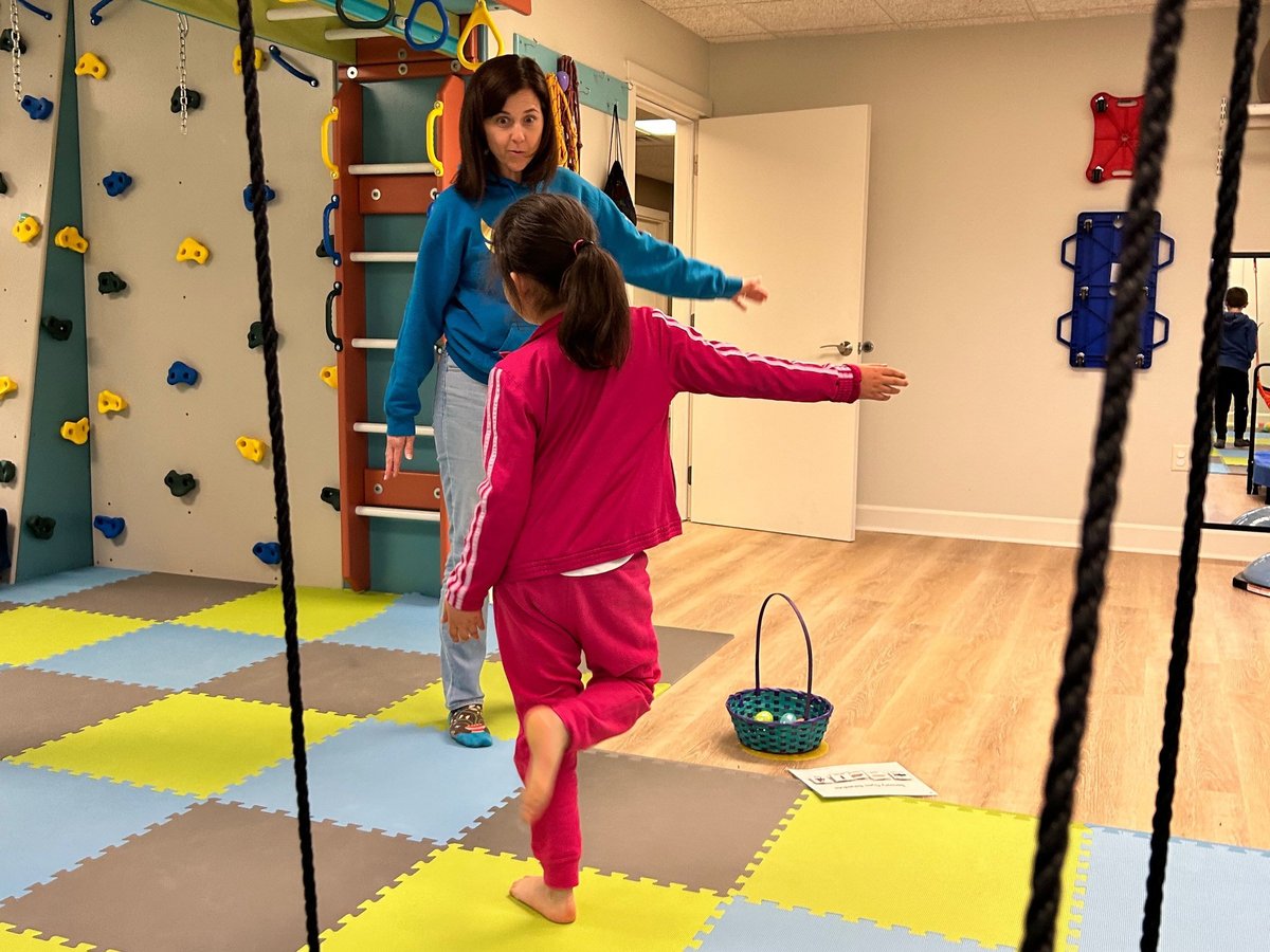 Child balancing on one leg while a therapist watches in the sensory gym with climbing wall, foam mats and suspension ropes