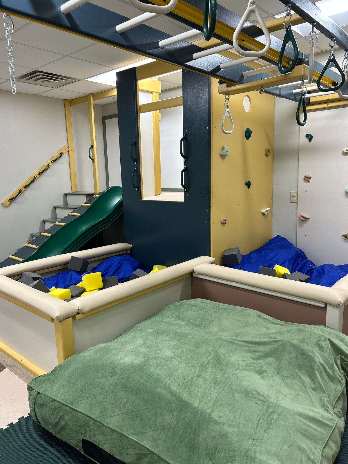 Indoor childrens play structure with climbing wall, slide, suspension rings, foam pit, and landing pads in the sensory gym at New Heights Therapy in Topeka, Kansas