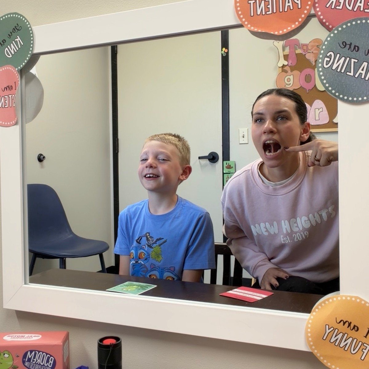 Two children smiling inside a classroom puppet theater window with decorative paper circles and signs on the wall behind them