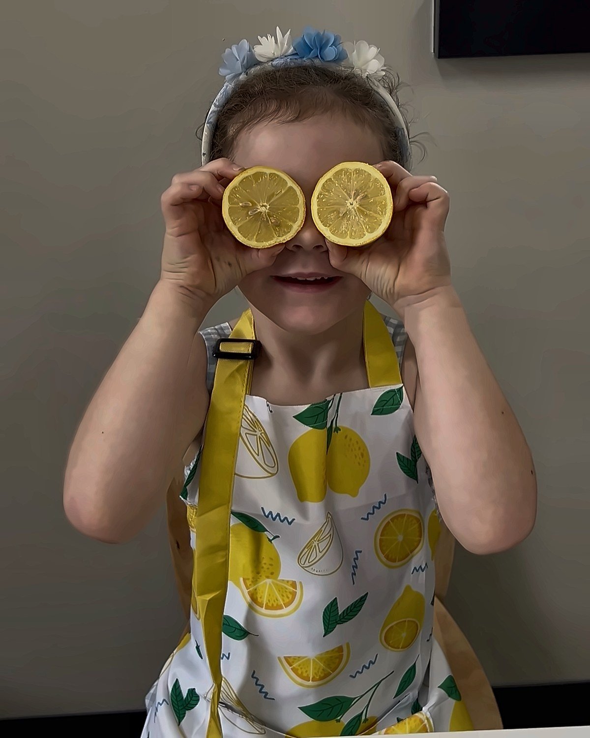 Young girl in a lemon-print apron holding lemon slices up to her eyes like glasses and smiling — playful food exploration during pediatric occupational and feeding therapy at New Heights Therapy in Kansas