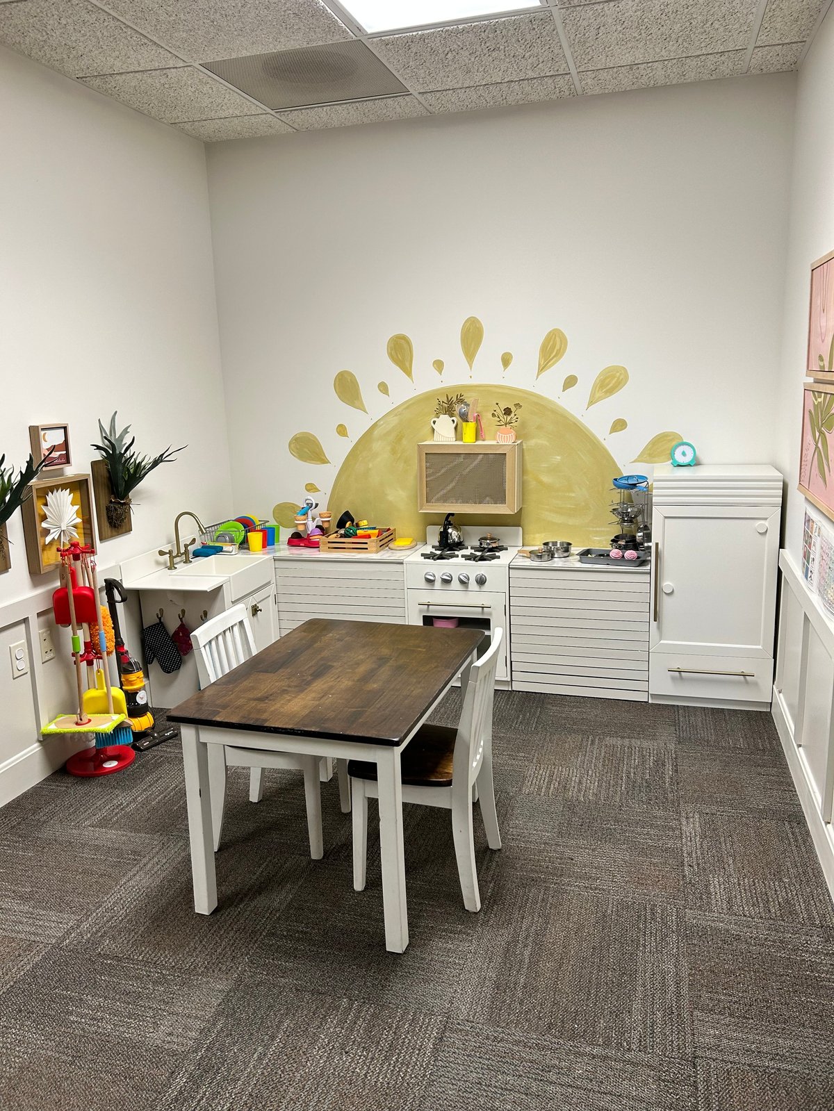 Children's play kitchen with white cabinetry, a wooden table and chairs, colorful toys and a large yellow sun mural