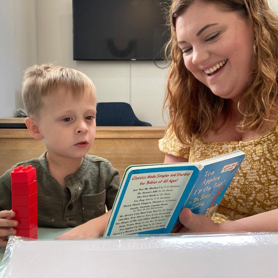 Pediatric speech therapist reading a Dr. Seuss book to a young boy with a red stacking toy during speech therapy at New Heights Therapy in Lawrence and Topeka, Kansas