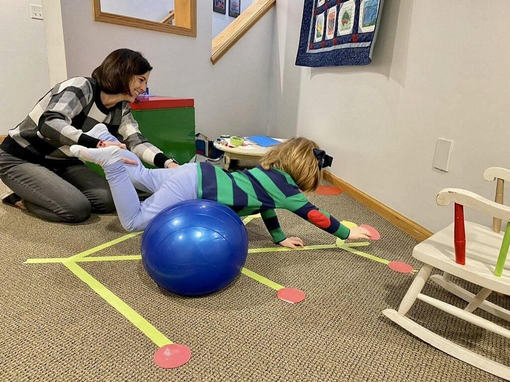 Occupational therapist supporting a child walking with hands on a blue therapy ball, a wheelbarrow-style balance exercise