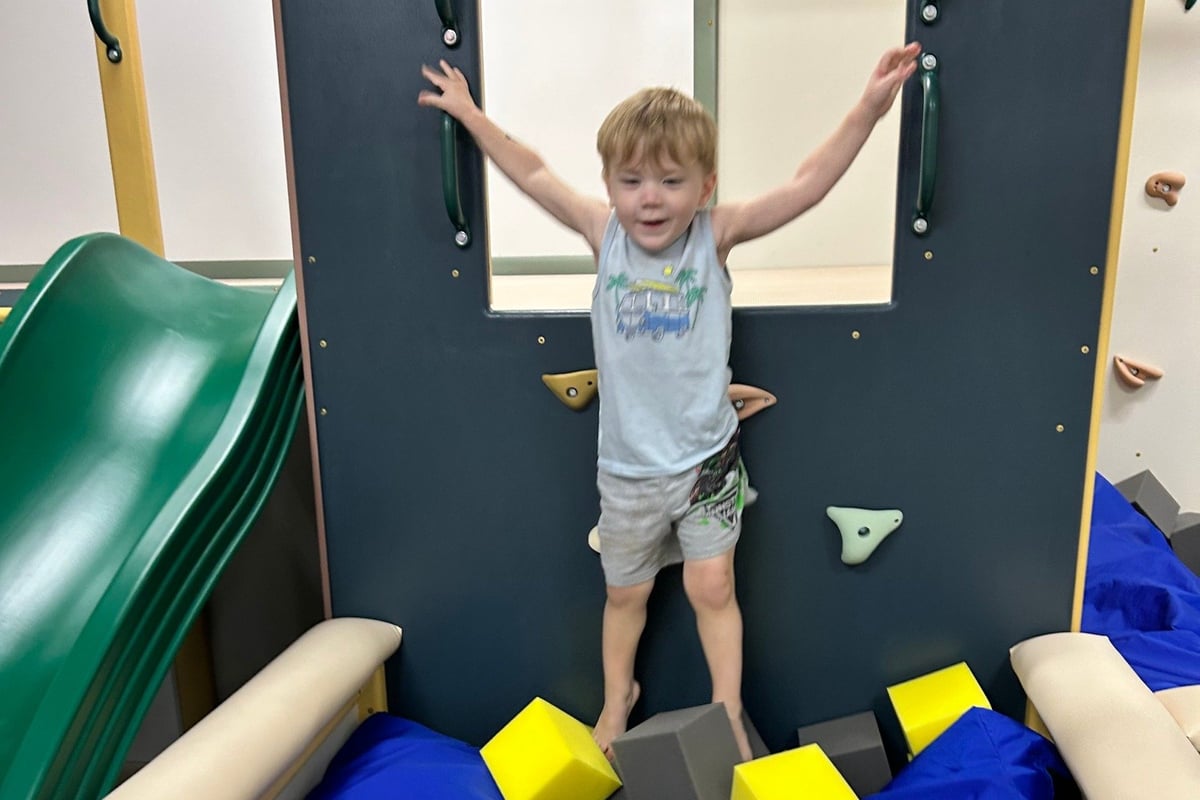 Young child with arms raised playing on indoor climbing wall with colorful foam blocks and holds.