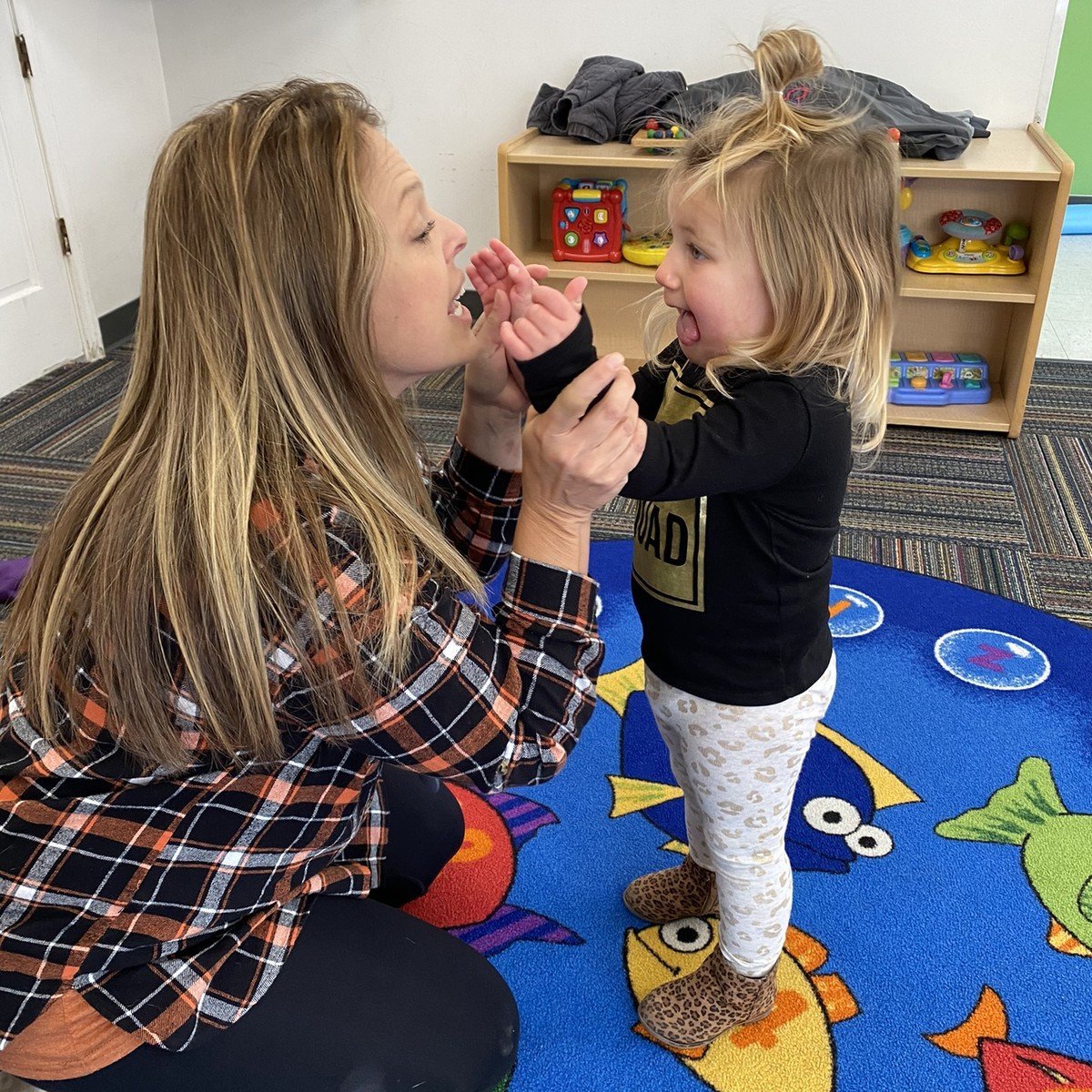 Woman and toddler playing with toy food in a colorful classroom setting with shelving and toys in background