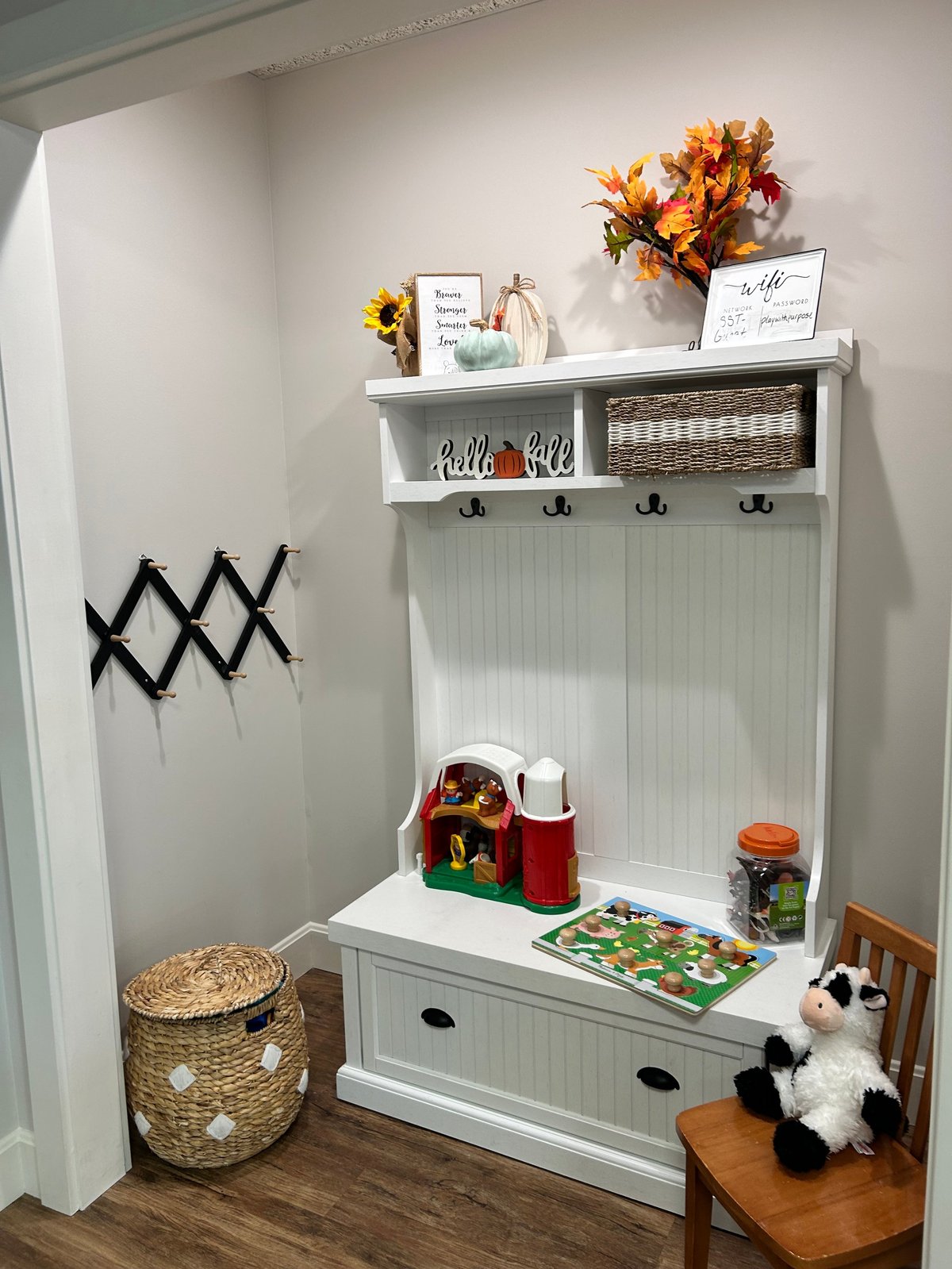 White entryway organization station with shelf, hooks, storage bench, decorative flowers, baskets, and childrens toys on wood flooring