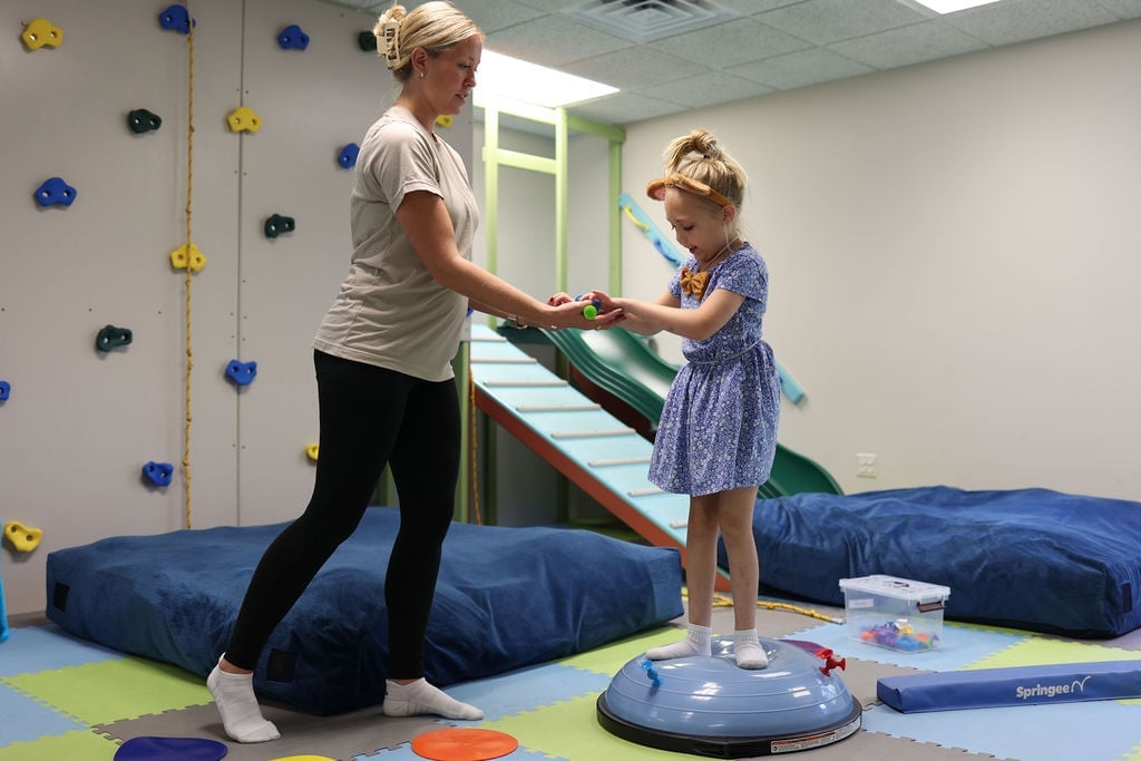 Pediatric physical therapist in Lawrence, KS steadying a young girl balancing on a blue wobble board in the New Heights Therapy sensory gym, with climbing wall and slide behind her