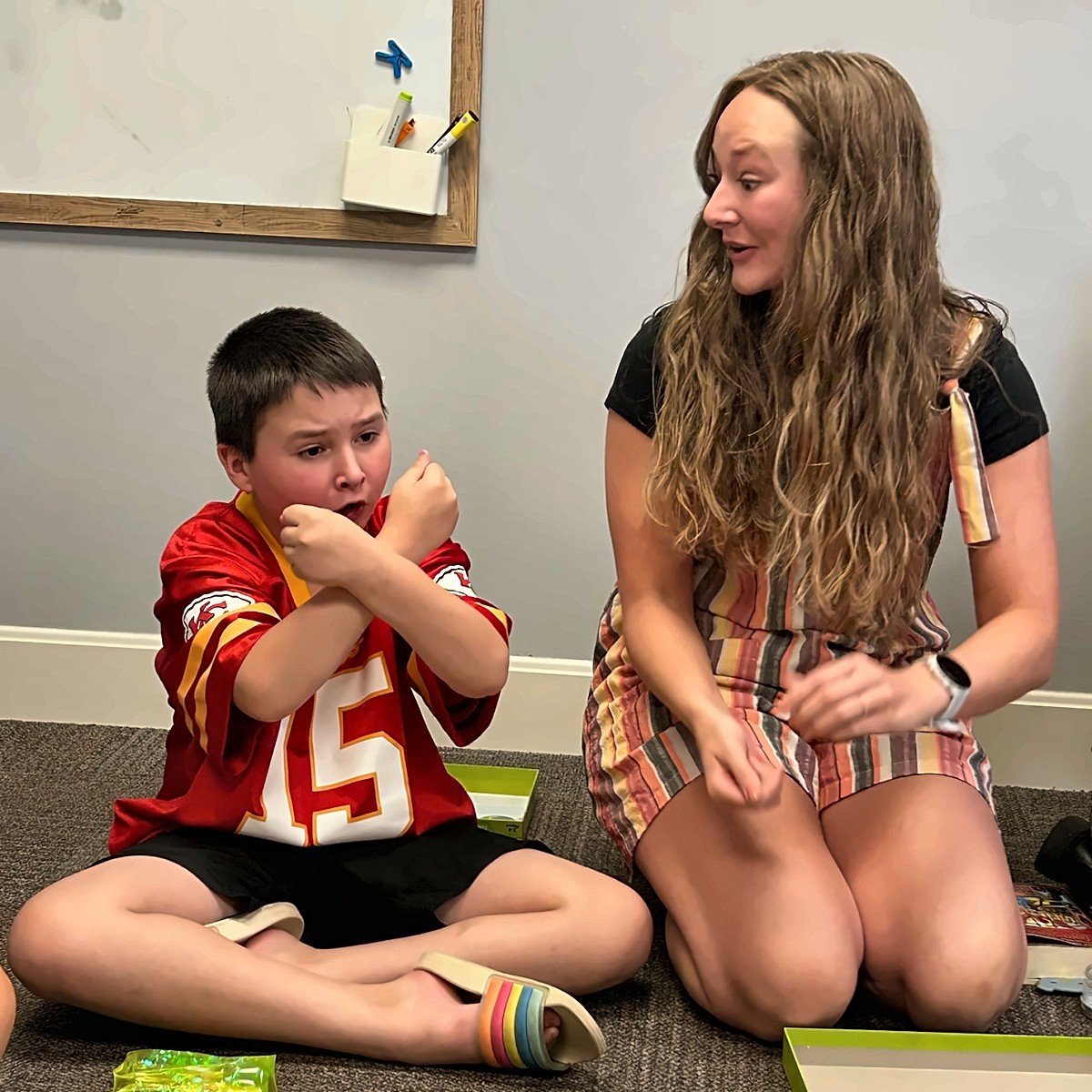 A young woman kneeling beside a young boy wearing a red sports jersey, engaging in conversation in an indoor classroom setting