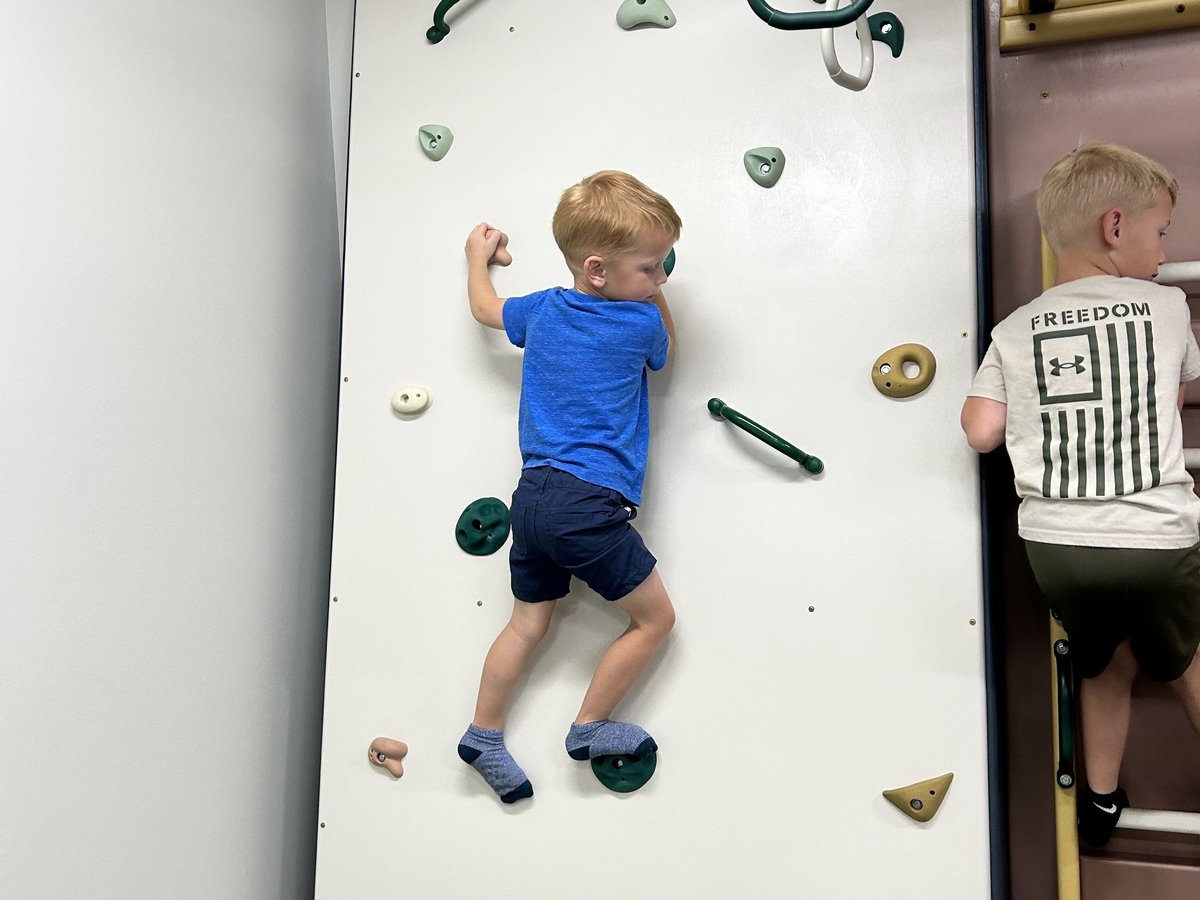 Young child in a blue shirt climbing the indoor climbing wall with colourful holds during an occupational therapy session in the sensory gym