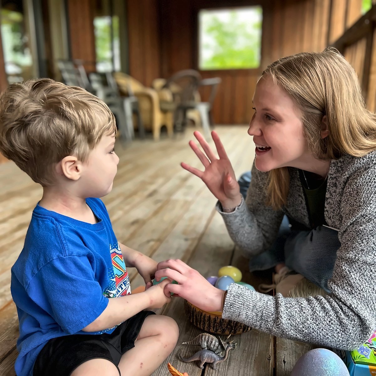 Woman in gray sweater smiling while interacting with young boy in blue shirt indoors on wooden floor