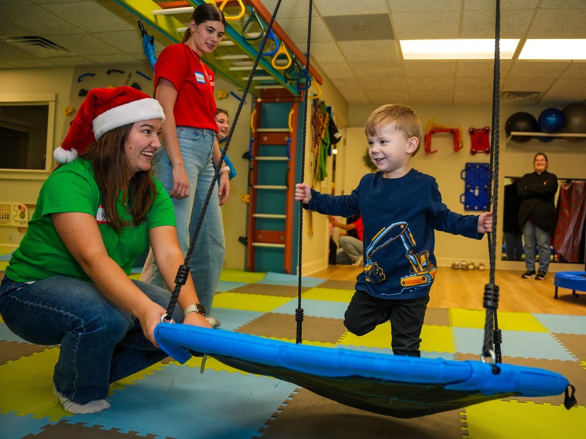 Therapist in a Santa hat helping a toddler balance on a blue platform swing during a holiday session in the colourful sensory gym