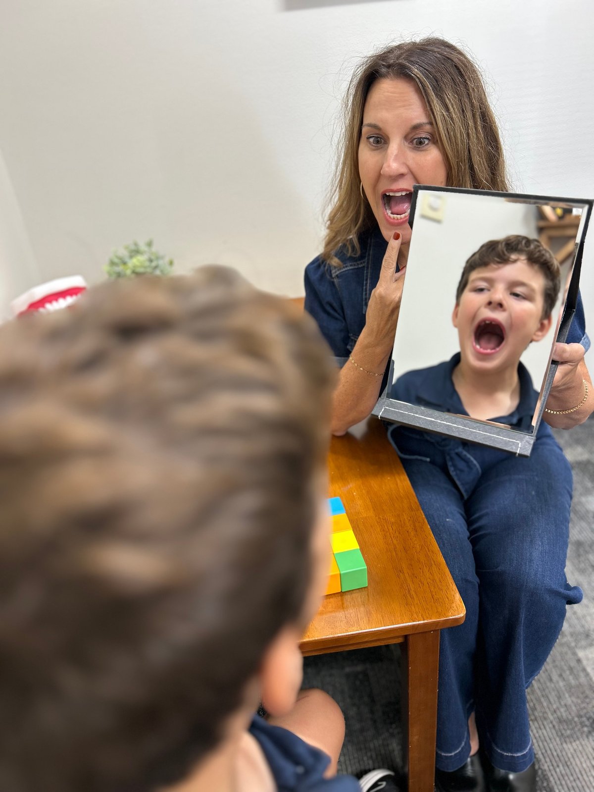 Orofacial myofunctional therapy in Lawrence and Topeka, KS — a therapist with her mouth wide open, pointing to her tongue, holding a mirror so a boy can copy the same tongue and jaw movement back to her